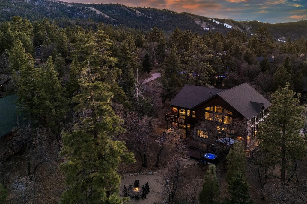 Aerial sunset view of Big Sky mountain cabin surrounded by forest and glowing outdoor firepit.