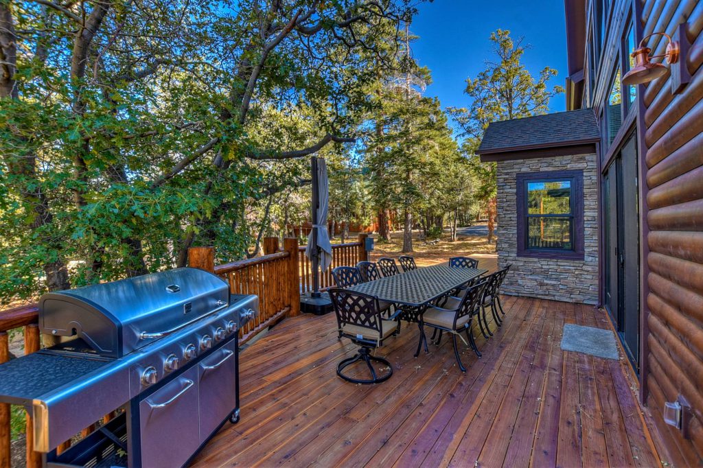 A spacious wooden deck with a large gas grill and an outdoor dining table with chairs is nestled among trees at a cabin in Big Bear.