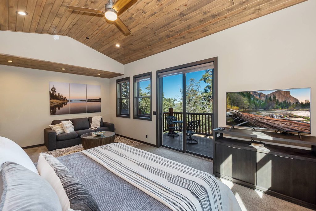 The interior of a master suite at Big Bear is shown, with a bed in the foreground, a small seating area, a large television, and sliding glass doors that lead to an outdoor deck.