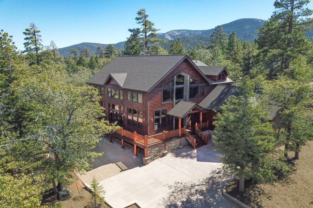 A large, multi-story wooden cabin with a modern design is shown in an elevated view, surrounded by pine trees with a mountain in the distance at Big Bear.