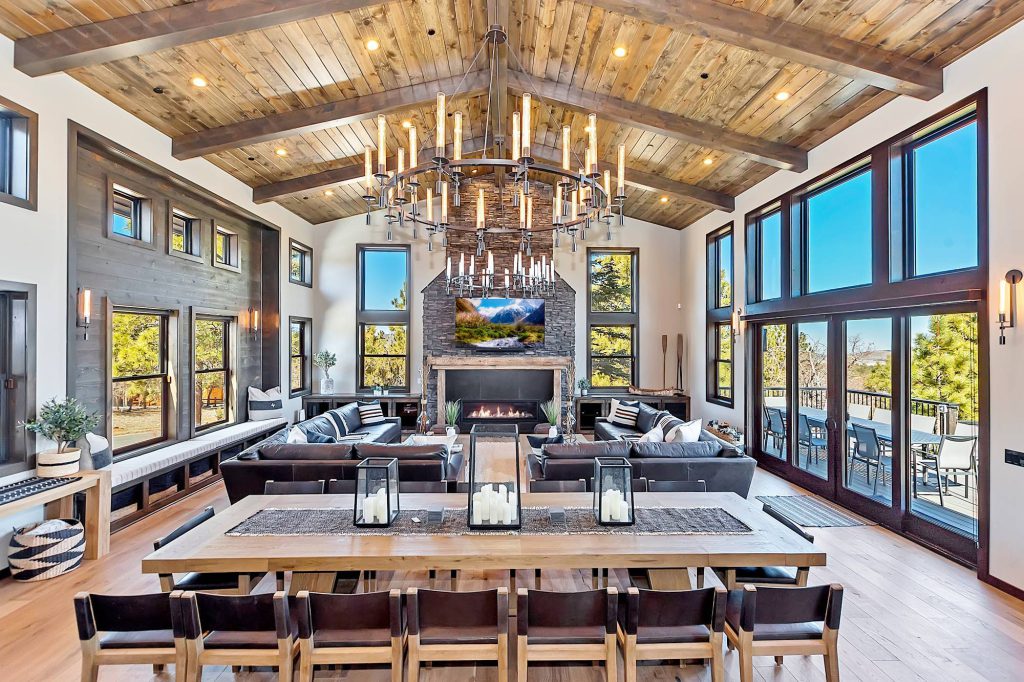 An interior view of a modern living and dining room with a large chandelier, high wood-paneled ceiling, and floor-to-ceiling windows.
