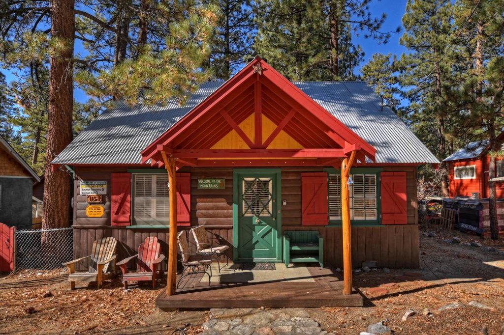 Charming rustic mountain cabin exterior with red and green accents, front porch seating, and forest surroundings