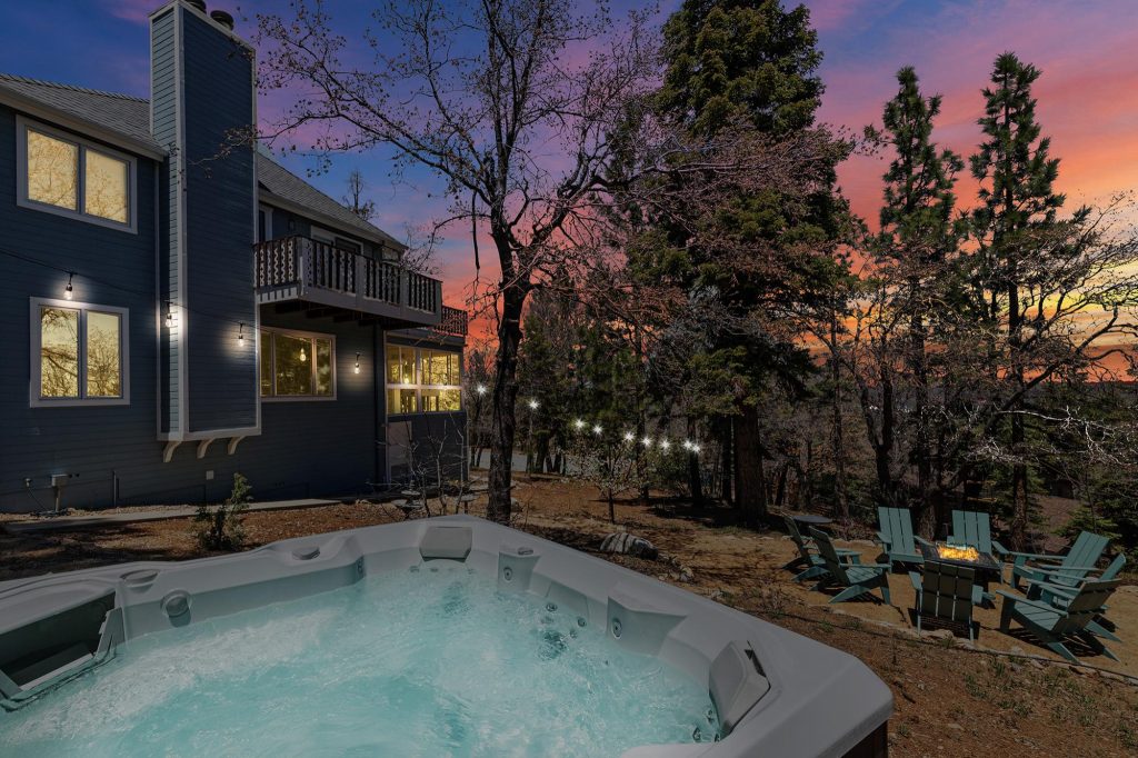 Backyard view with a hot tub in the foreground, outdoor seating around a fire pit, tall trees surrounding a blue house at sunset
