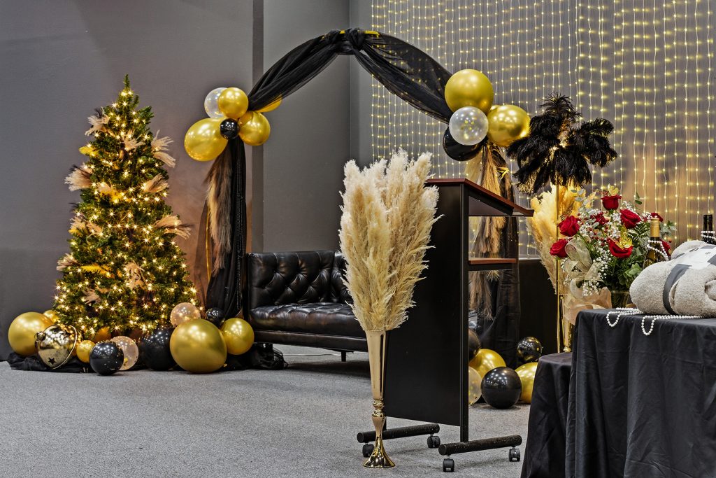 Event stage setup with black and gold balloon arch, decorative lights, and lounge seating