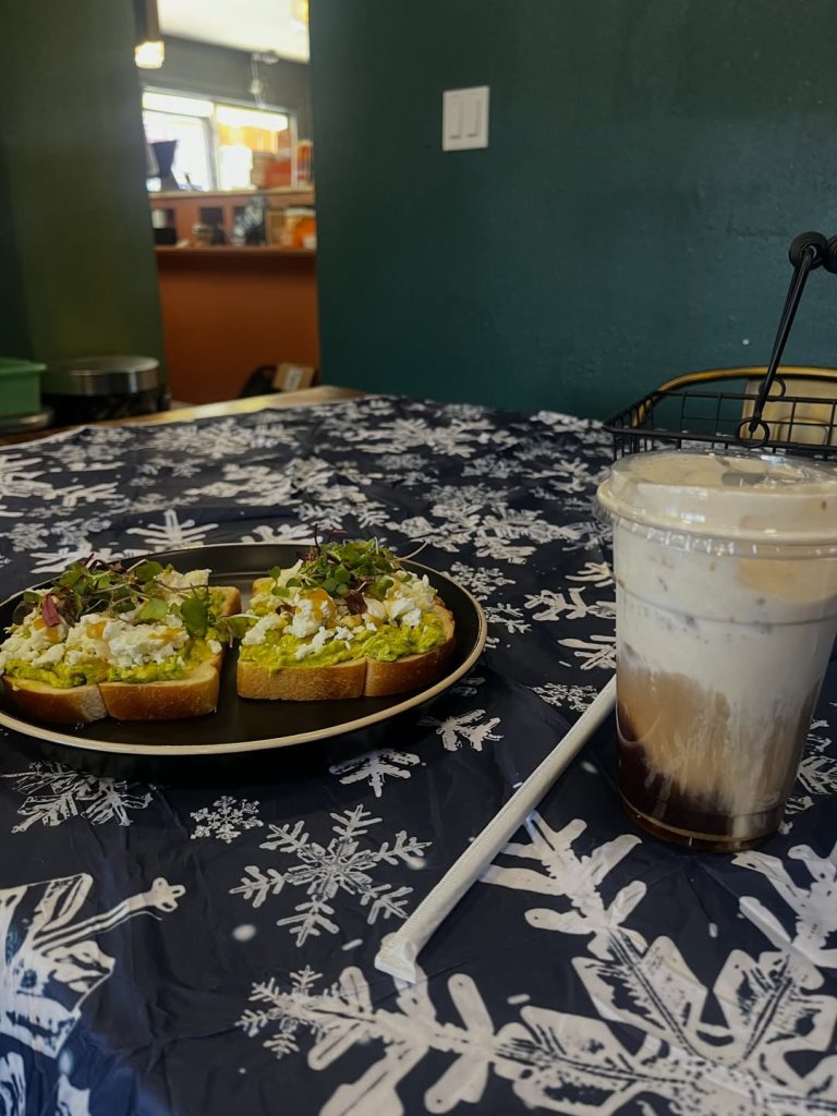 Plate with avocado toast topped with cheese and microgreens, alongside a cold iced coffee on a snowflake-patterned tablecloth.