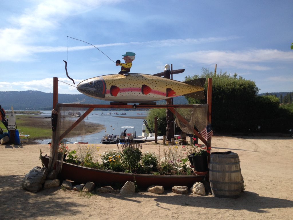 A large, colorful fish sculpture with a cartoon fisherman on top, serving as a sign near Big Bear Lake.