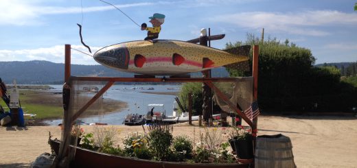 A large, colorful fish sculpture with a cartoon fisherman on top, serving as a sign near Big Bear Lake.