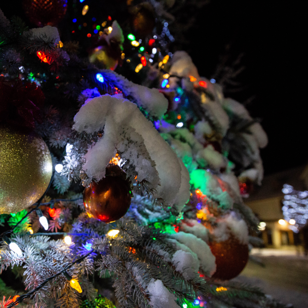 A close-up shot of an outdoor Christmas tree adorned with colorful lights and golden ornaments, heavily covered in white snow, set against a dark night background, celebrating Christmas at Big Bear.