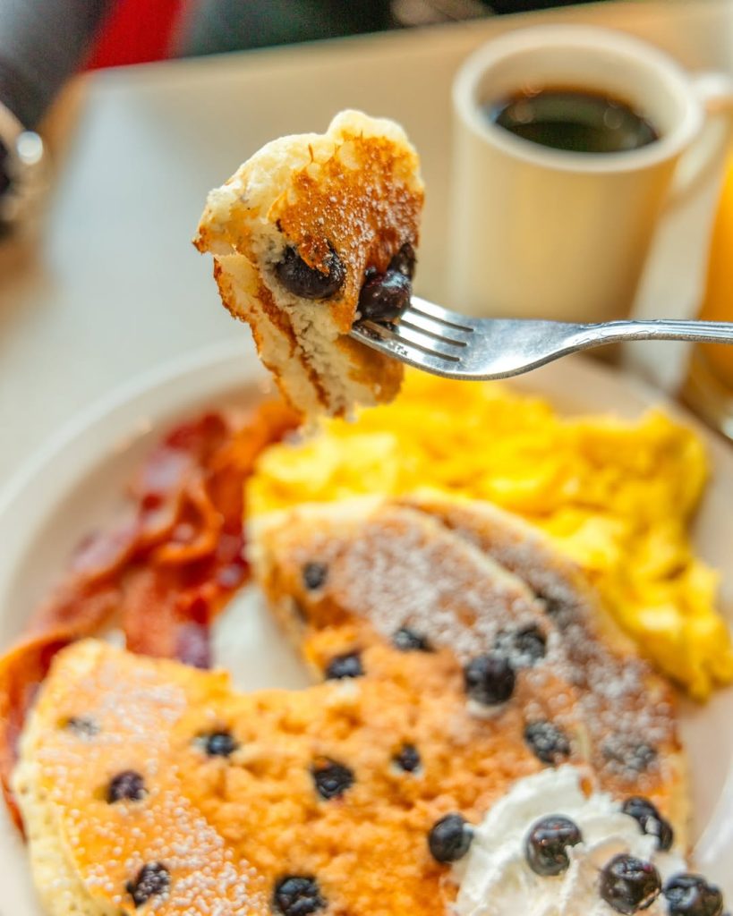 Close-up of blueberry pancakes served with eggs, bacon, and coffee at a breakfast table.