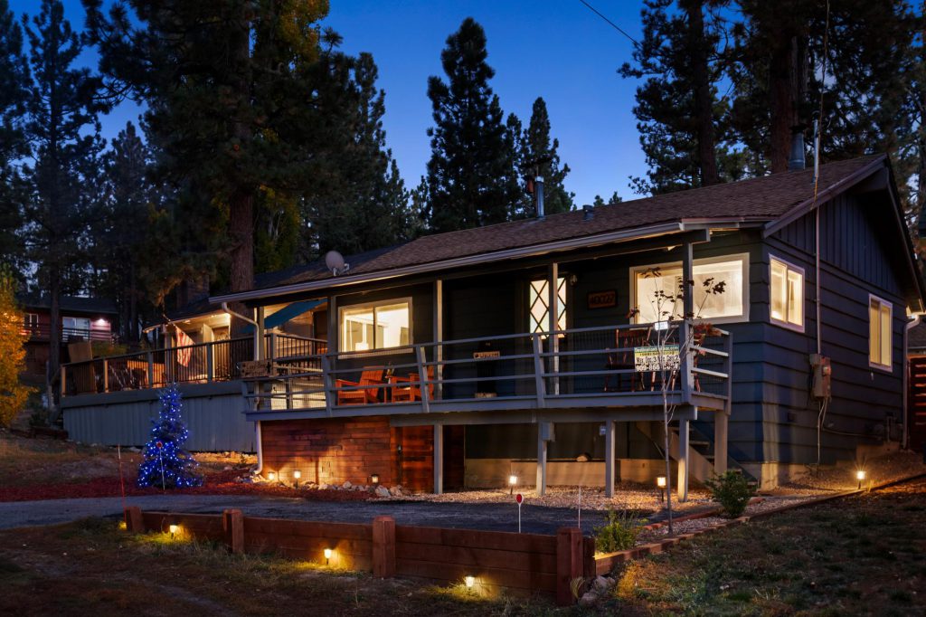 A beautiful, single-story dark gray cabin with a deck, a Christmas tree, and a lighted walkway is nestled among trees at dusk in Big Bear.