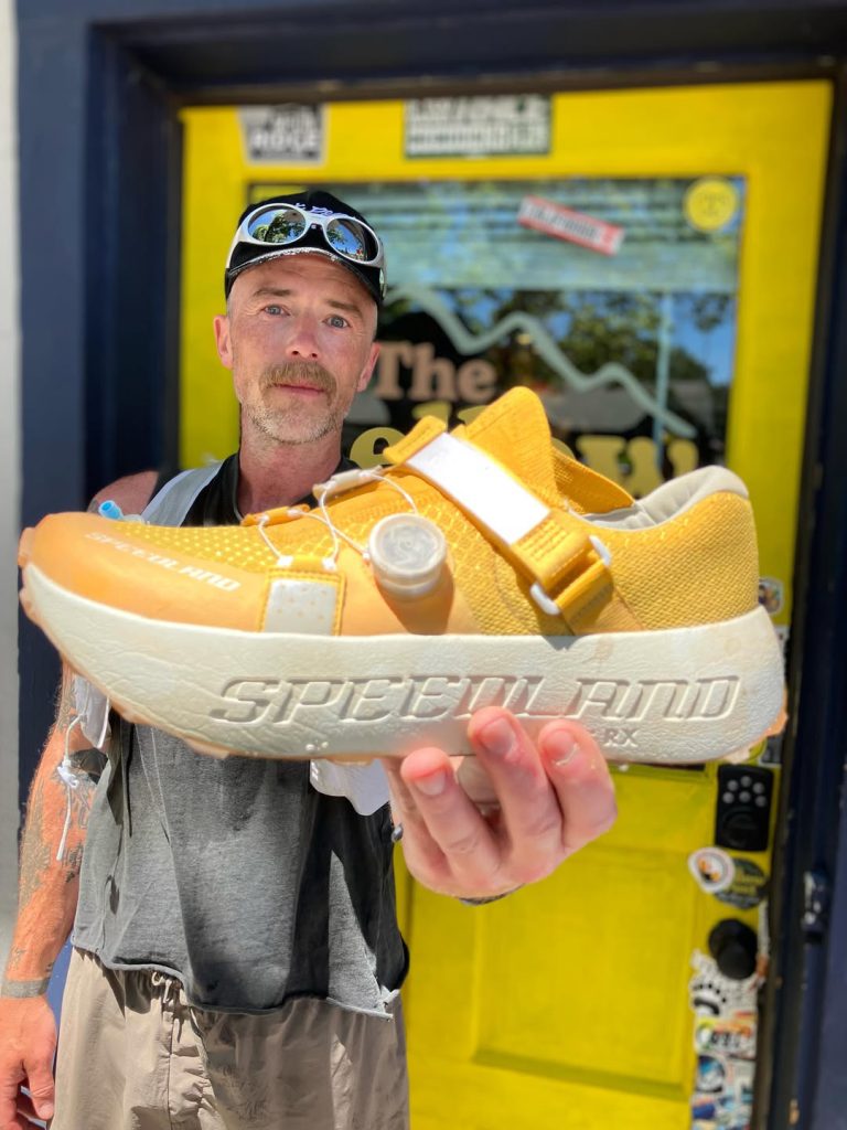 Man holding a bright yellow Specialized cycling shoe toward the camera in front of a shop entrance with a yellow door background.