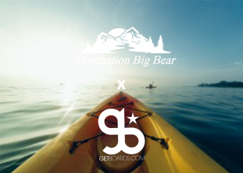 The point of view from a yellow kayak on a calm lake with mountains in the background, with the logo for "Destination Big Bear" and "GetBoards.com" overlaid on the image.