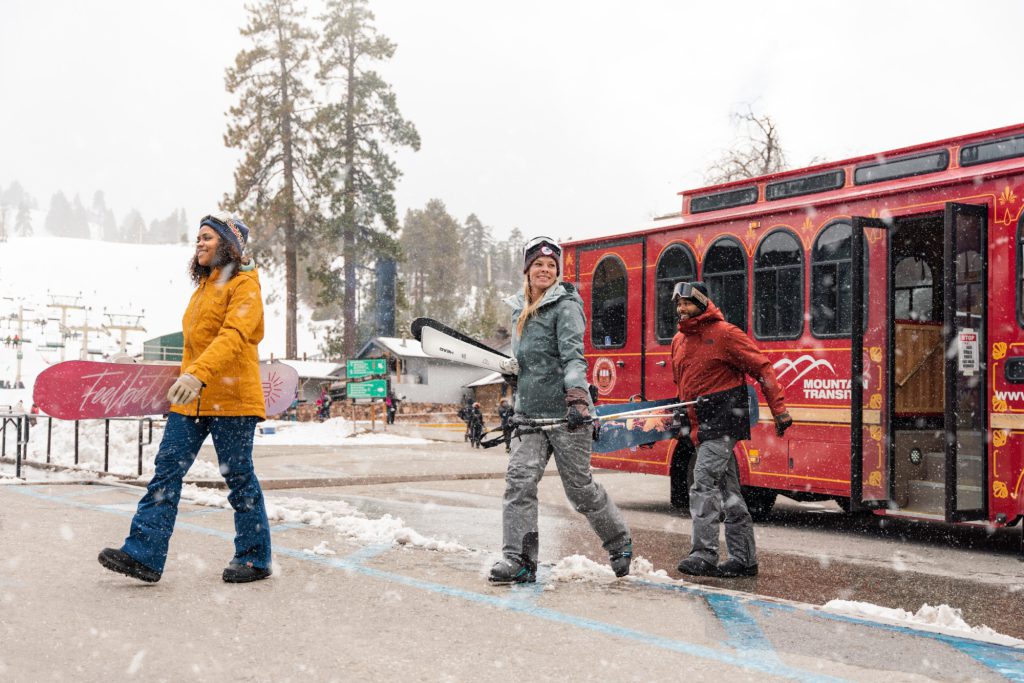 Three friends walking with snowboards and skis near a red trolley in a snowy Big Bear setting.