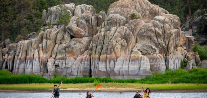 A group of four people paddleboarding on Big Bear Lake, with a massive pile of striated boulders and a lush, green treeline in the background.