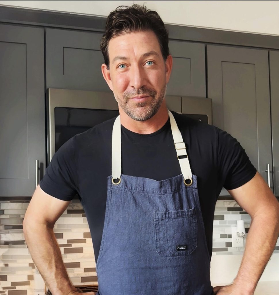 Dawson the Cook, wearing a blue apron, stands in front of a kitchen, ready to prepare meals in Big Bear.