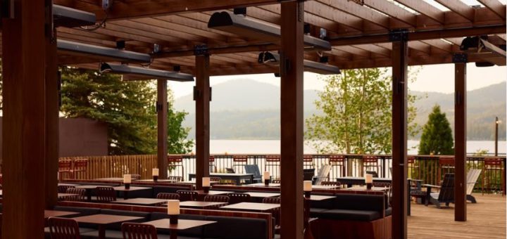 An outdoor patio at Big Bear with wooden tables, benches, and overhead heaters, overlooking a lake surrounded by mountains and trees.