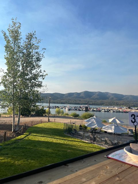 A view of Big Bear Lake from a patio, showing the water with boats, a green lawn, mountains in the distance, and white umbrellas.