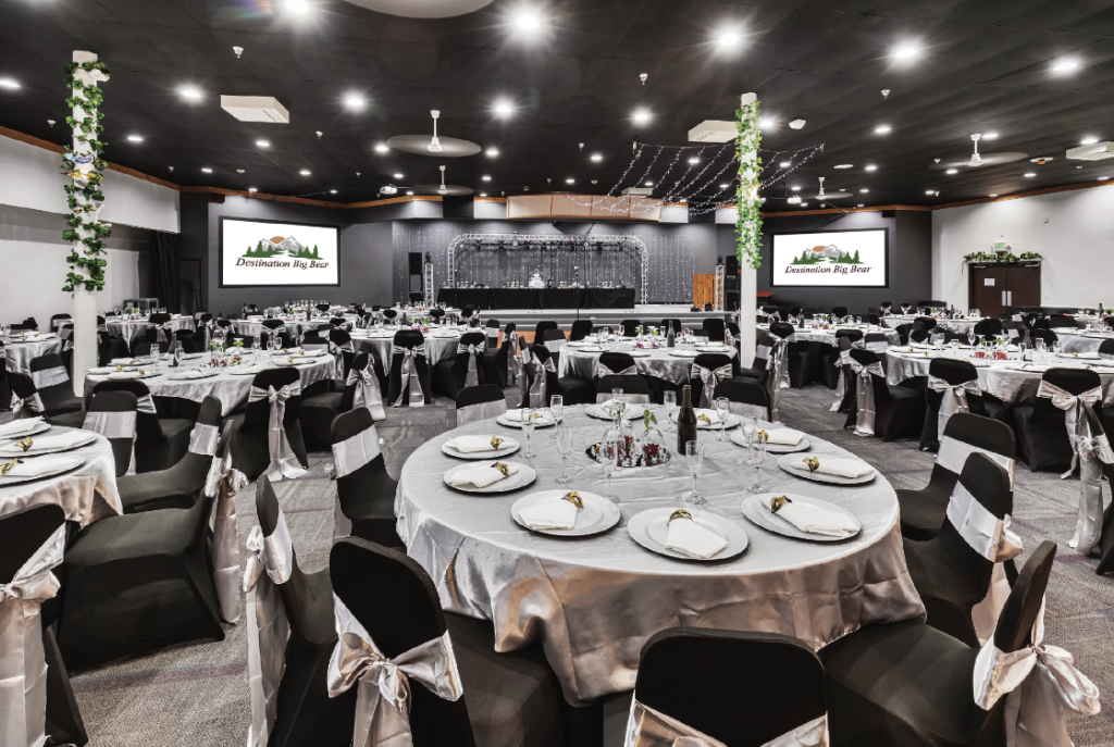 A large, elegant event hall in Big Bear is set up for a formal dinner with multiple round tables covered in white tablecloths and black chairs.