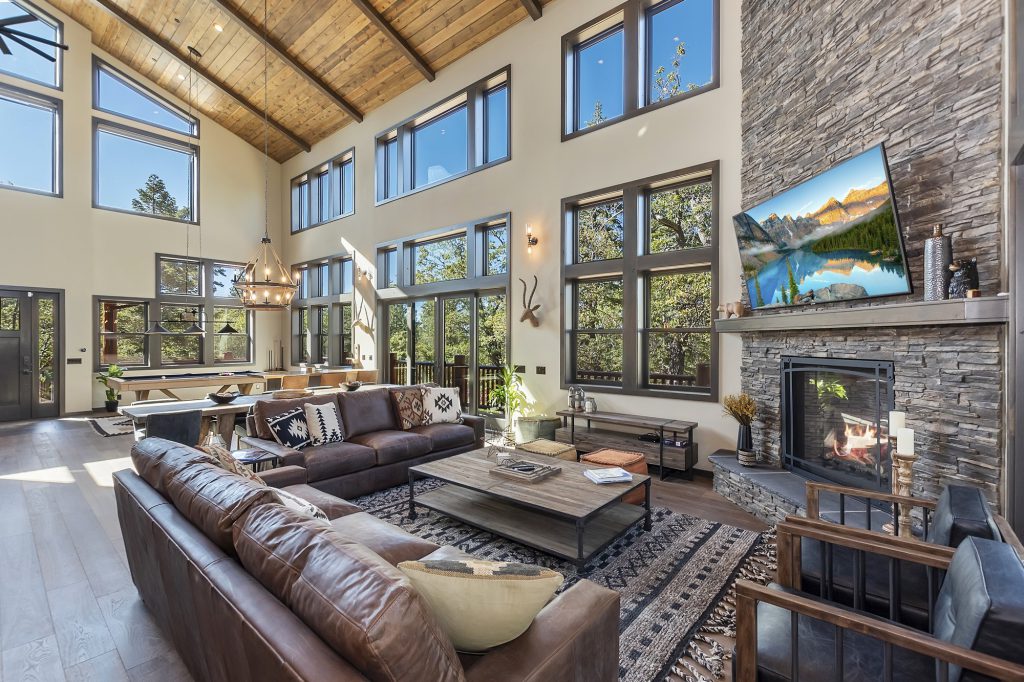 A large, high-ceilinged great room in a Big Bear home, with a stone fireplace, leather sofas, a pool table in the background, and multiple large windows looking out onto a wooded area.