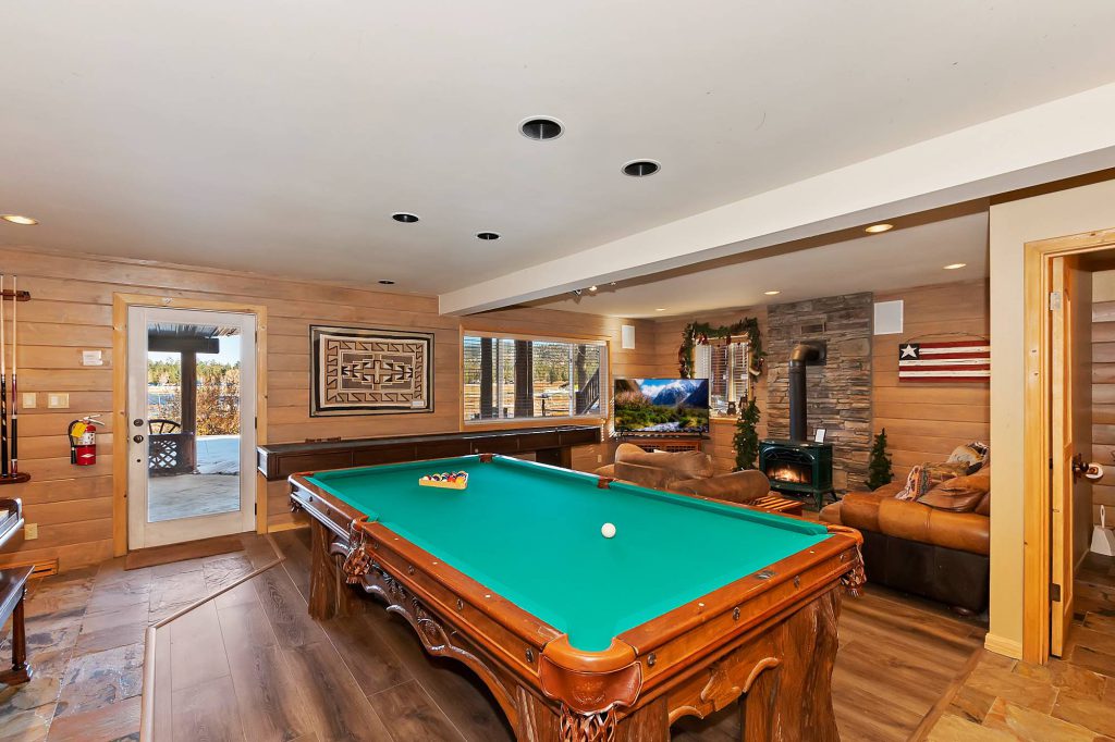 The game room of a Big Bear cabin is shown, featuring a green felt pool table, a foosball table, wood paneling on the walls, and a stone fireplace with a view of a snowy outdoor area.