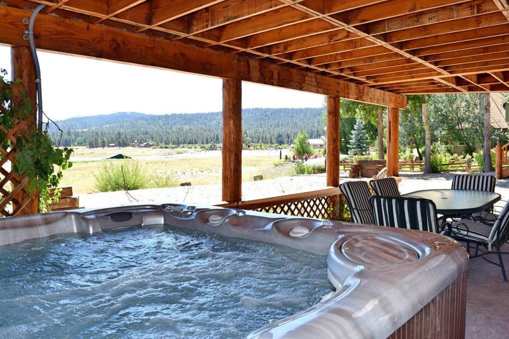 A hot tub on a covered patio at a Big Bear cabin with an outdoor dining table and a view of an open field with trees and mountains in the background.