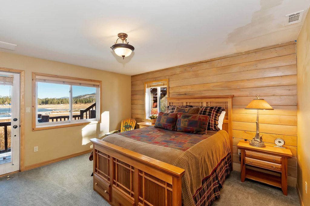 A bedroom with a wooden bed frame, a patterned comforter, and wood-paneled walls, featuring a large window and a sliding glass door that both offer a view of Big Bear Lake.