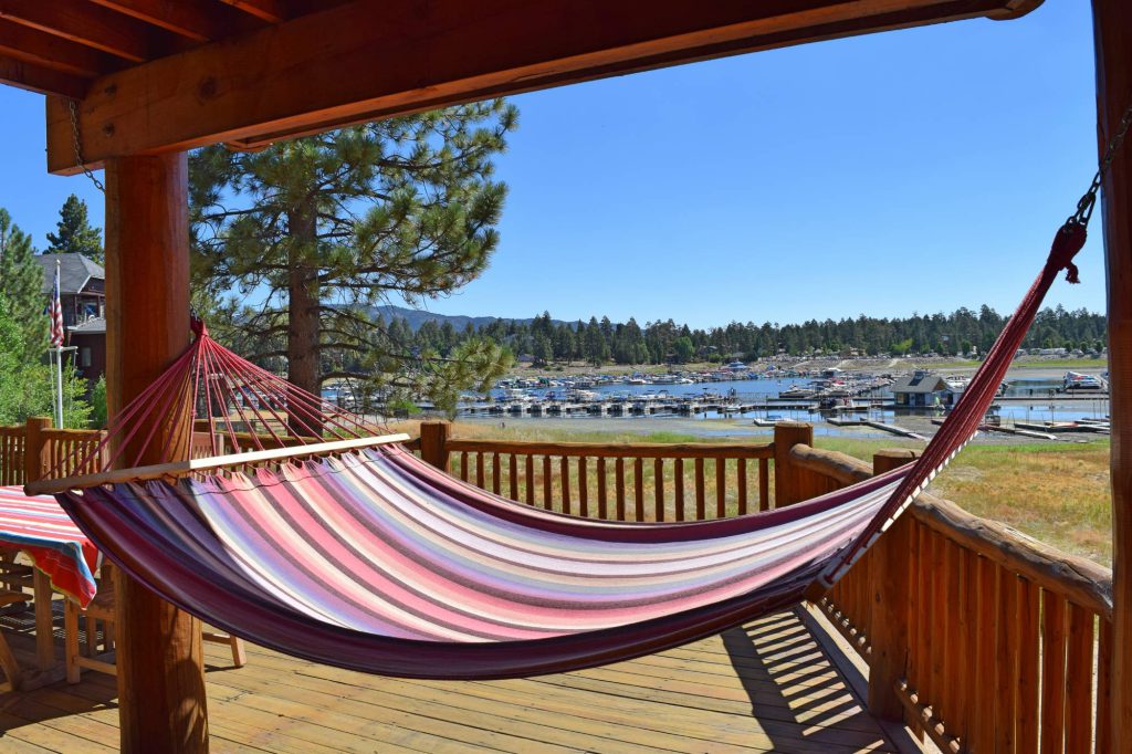 A colorful striped hammock hangs on a wooden deck overlooking a marina on Big Bear Lake, with boats docked and mountains in the background under a clear blue sky.