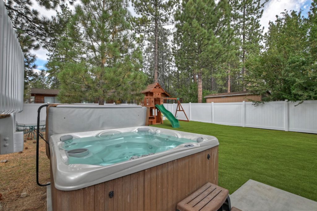 Backyard hot tub with fenced lawn and children's playset surrounded by tall pine trees