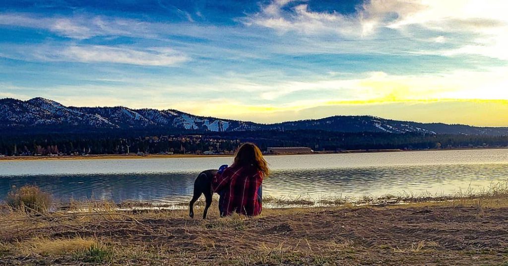 A beautiful scene at Big Bear shows a person and their dog sitting by the lake, with mountains in the background under a colorful sky.