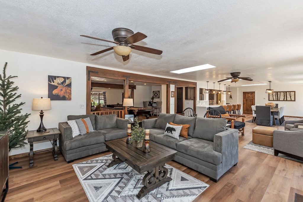 An open-concept cabin living area in Big Bear with a gray sectional sofa, rustic wooden furniture, and a dining area and game room visible in the background.