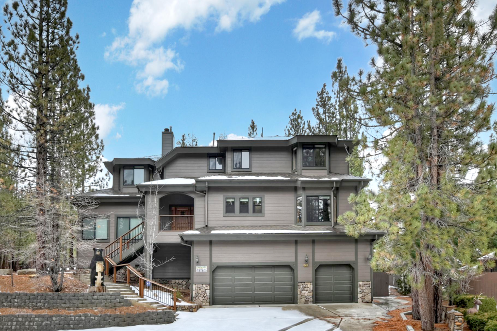 A large, modern, three-story cabin with a two-car garage is surrounded by pine trees on a snowy day in Big Bear.