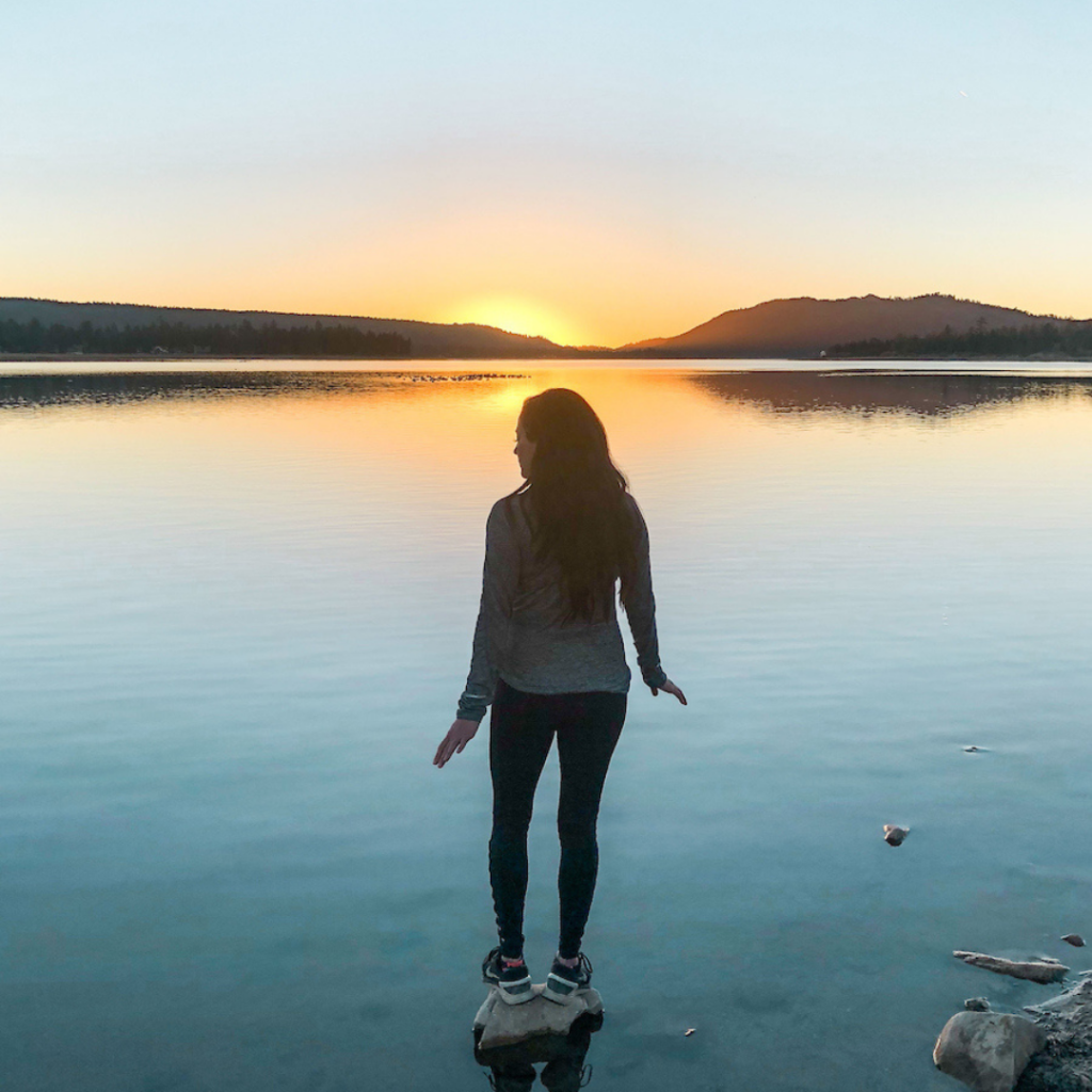 A woman stands on a rock at the edge of the water at Big Bear, looking out at a stunning sunset over the lake and mountains.
