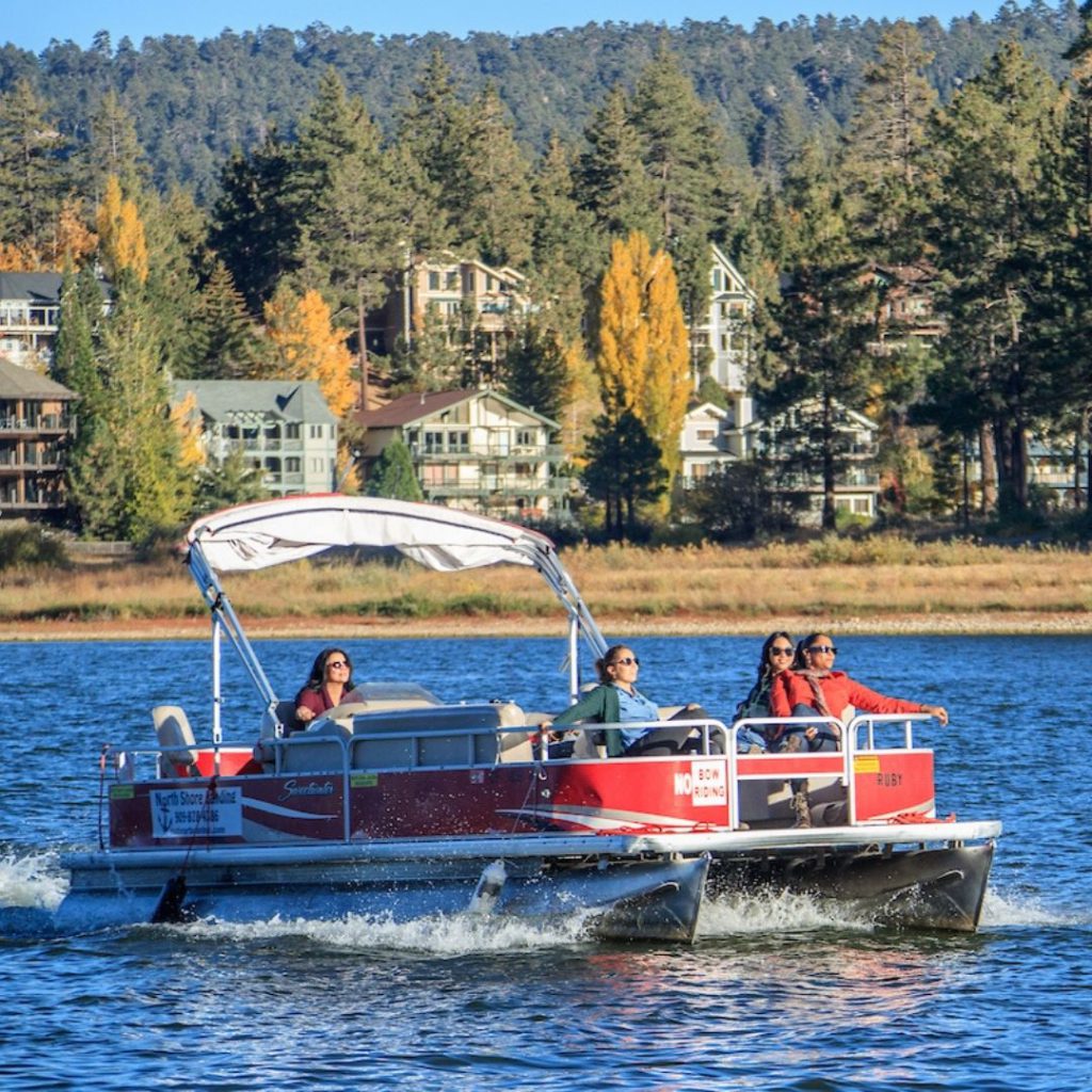 Red and white pontoon boat with people enjoying a relaxing outing at Big Bear Lake, surrounded by scenic forested hills.