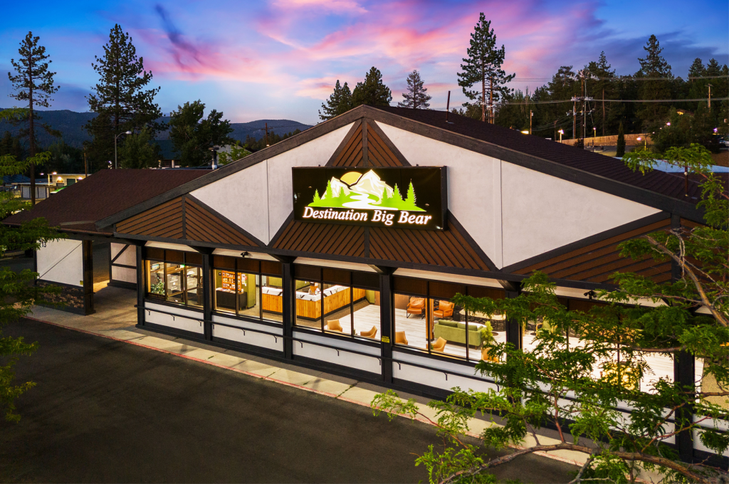 The front of the Destination Big Bear building is illuminated at dusk, with its logo visible and the sky painted with beautiful sunset colors.