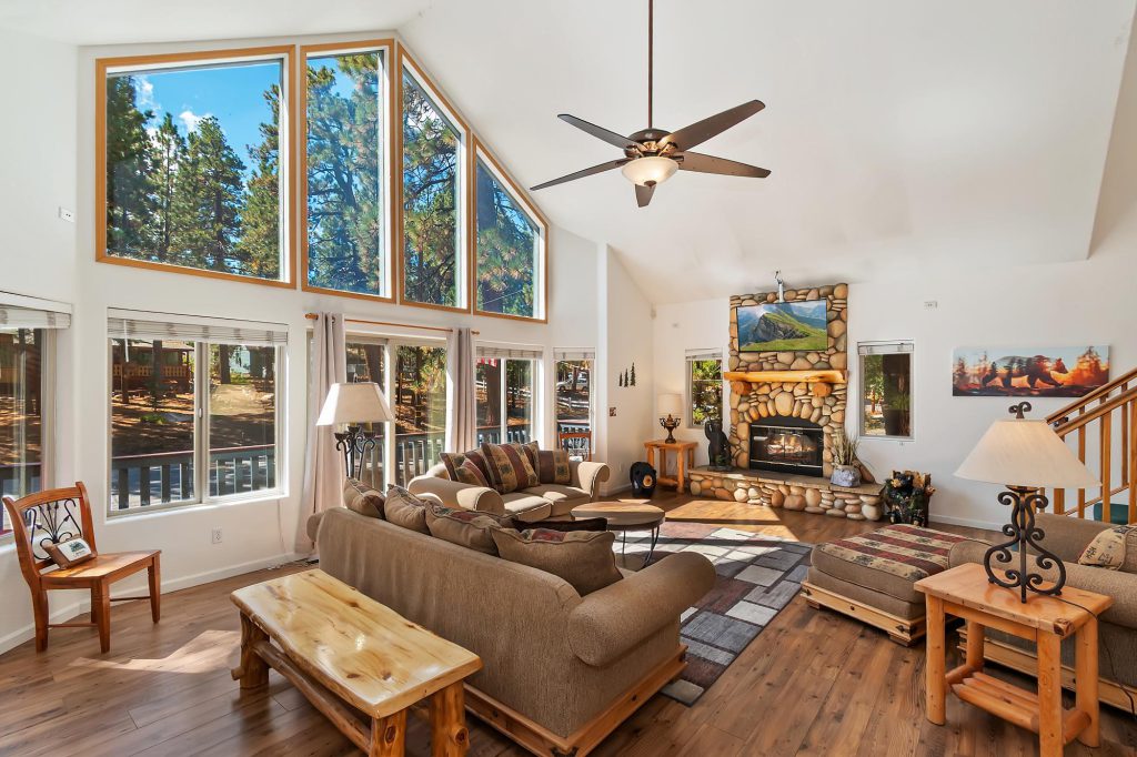 Living Room with a sofa and loveseat in front of a 15' high ceiling with a full wall of windows behind the loveseat. A stone fireplace on a far wall. Outside the windows is a mountain setting.