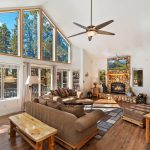 Living Room with a sofa and loveseat in front of a 15' high ceiling with a full wall of windows behind the loveseat. A stone fireplace on a far wall. Outside the windows is a mountain setting.