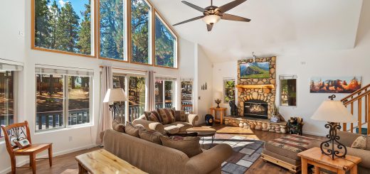 Living Room with a sofa and loveseat in front of a 15' high ceiling with a full wall of windows behind the loveseat. A stone fireplace on a far wall. Outside the windows is a mountain setting.