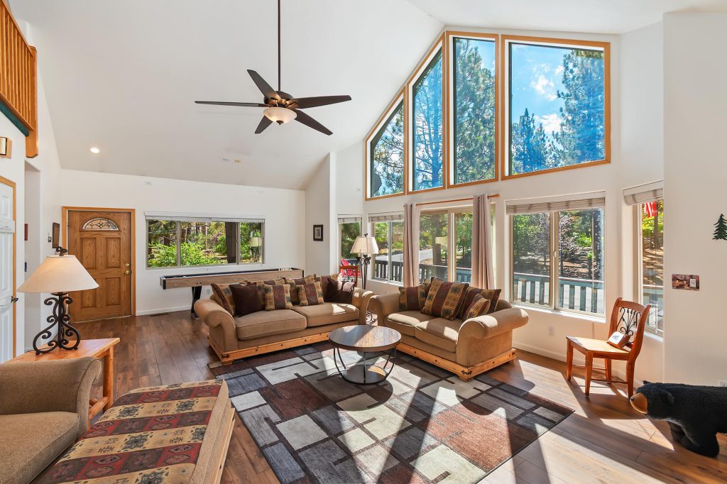Living Room with a sofa and loveseat in front of a 15' high ceiling with a full wall of windows behind the loveseat. Outside the windows is a mountain setting.