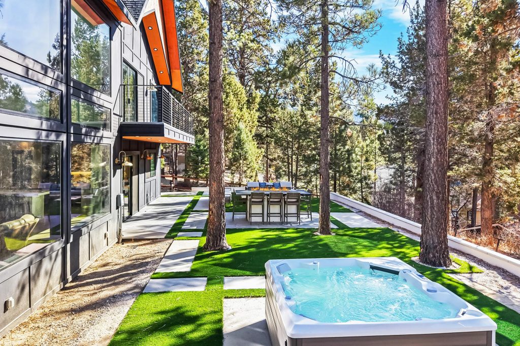 Luxury cabin backyard in Big Bear featuring a private hot tub, outdoor dining area, lush artificial turf, and tall pine trees in a peaceful forest setting.