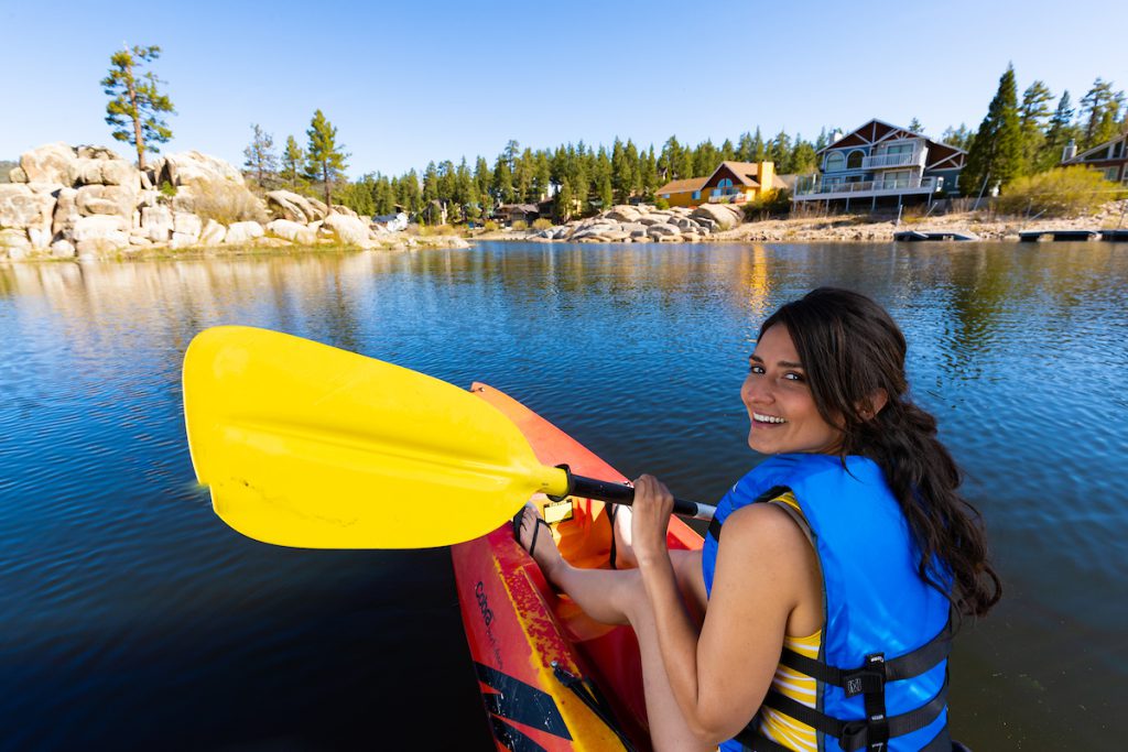 A smiling woman in a life vest is paddling a kayak on Big Bear Lake, with houses and rocks visible on the shore in the background.