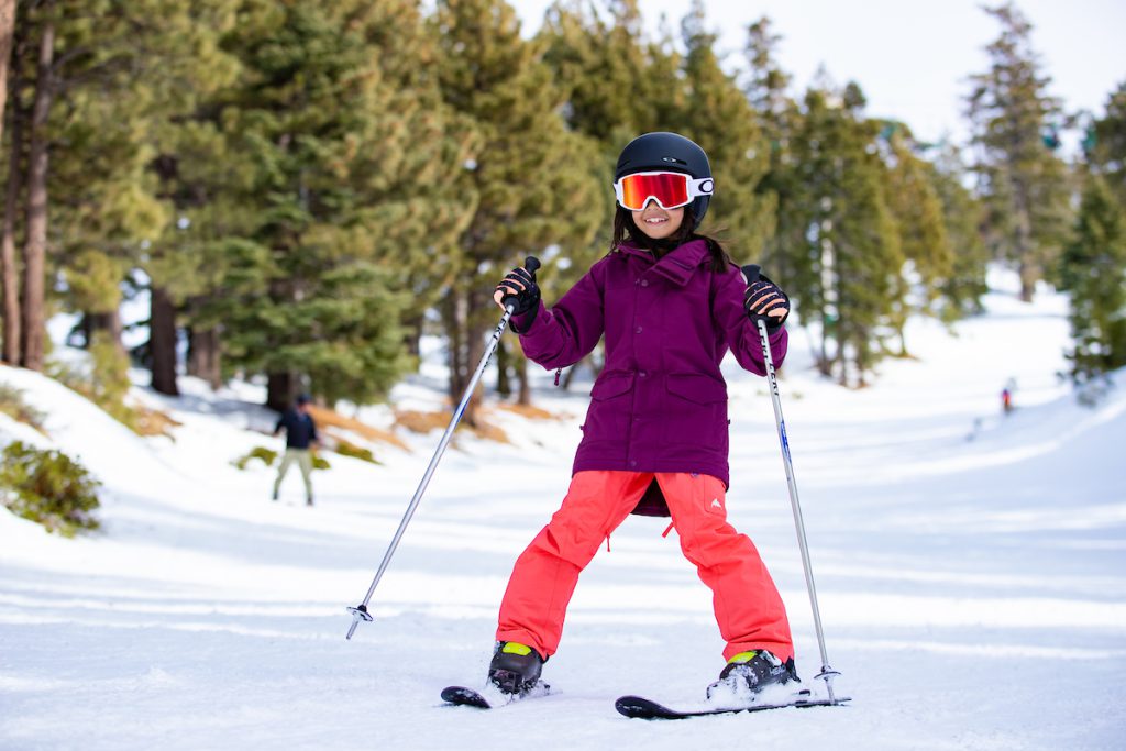 A child in a purple jacket and orange pants skis on a snowy slope at Big Bear.