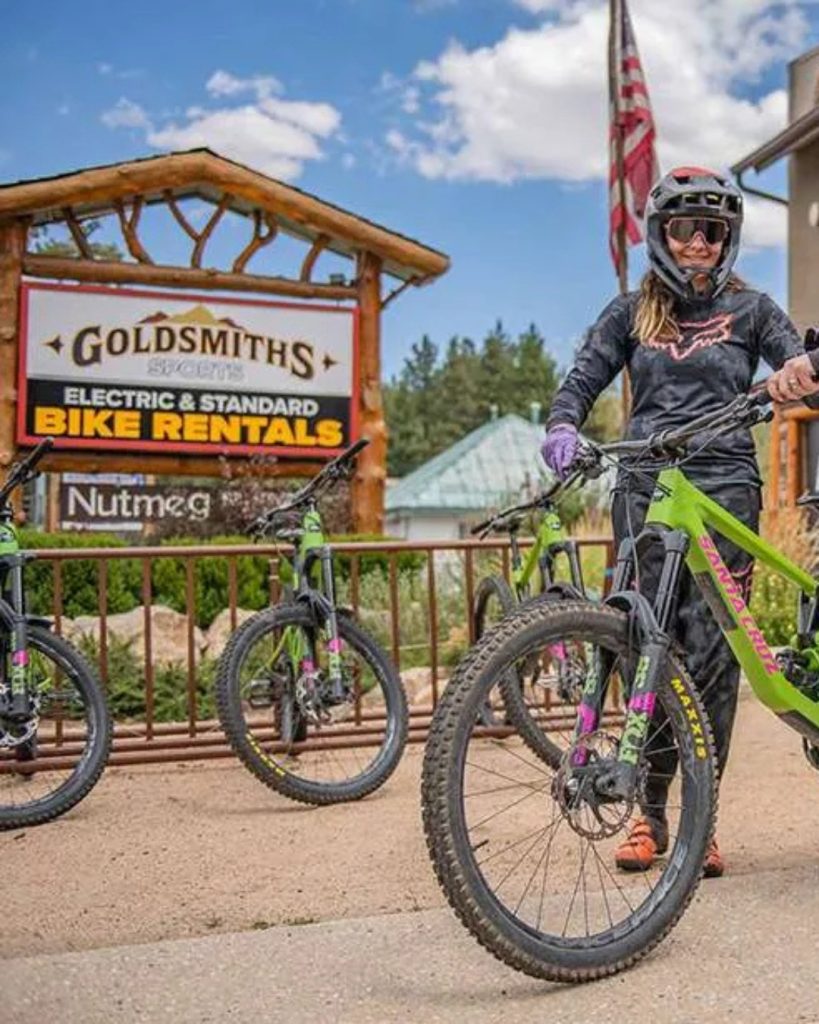A cyclist stands beside a mountain bike outside Goldsmiths Sports bike rental shop in Big Bear, with rental bikes and signage visible.