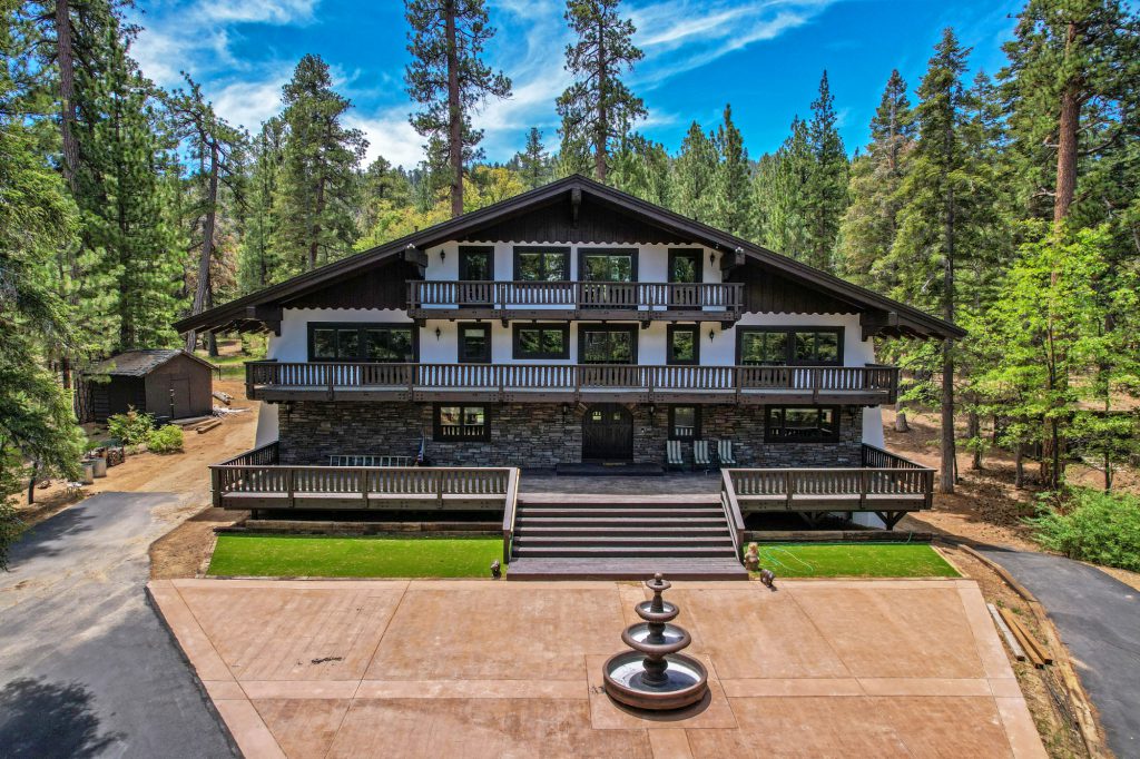 A large rustic lodge with a stone facade and a fountain is situated in a forest clearing in Big Bear.