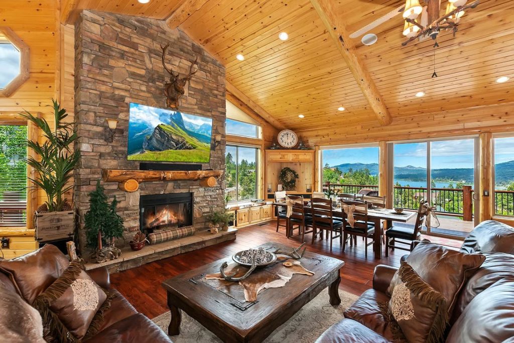 Spacious Big Bear lodge interior with a stone fireplace, wood-paneled ceiling, leather seating, dining area, and lake views