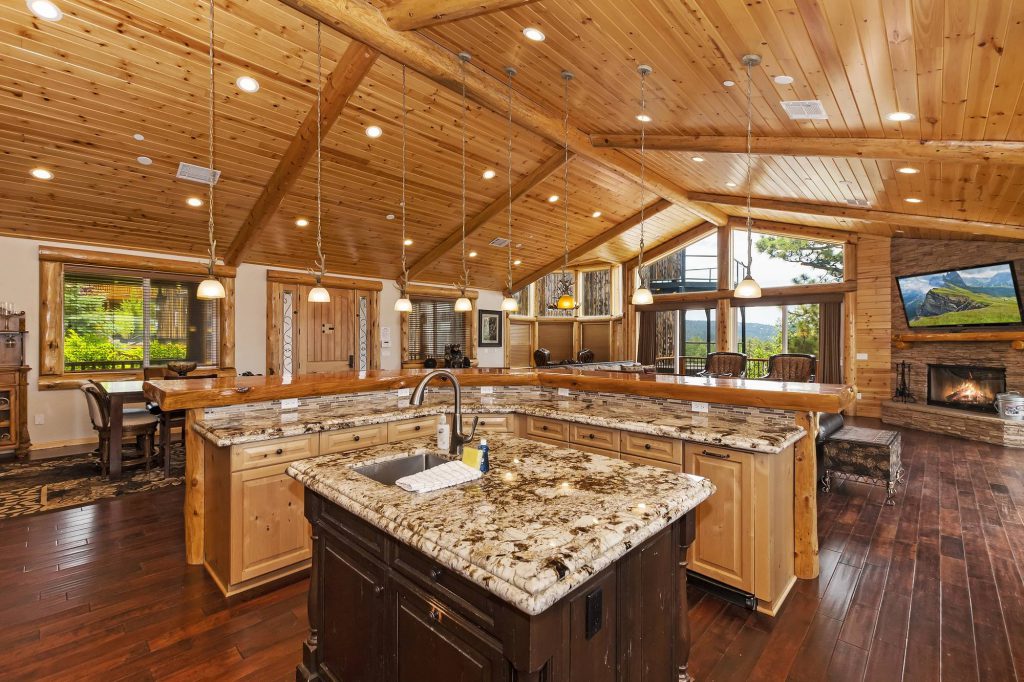 A modern kitchen with a large, square island with a sink in the center is visible, with dark wood floors, light wood ceilings, and a stone fireplace in the background in Big Bear.