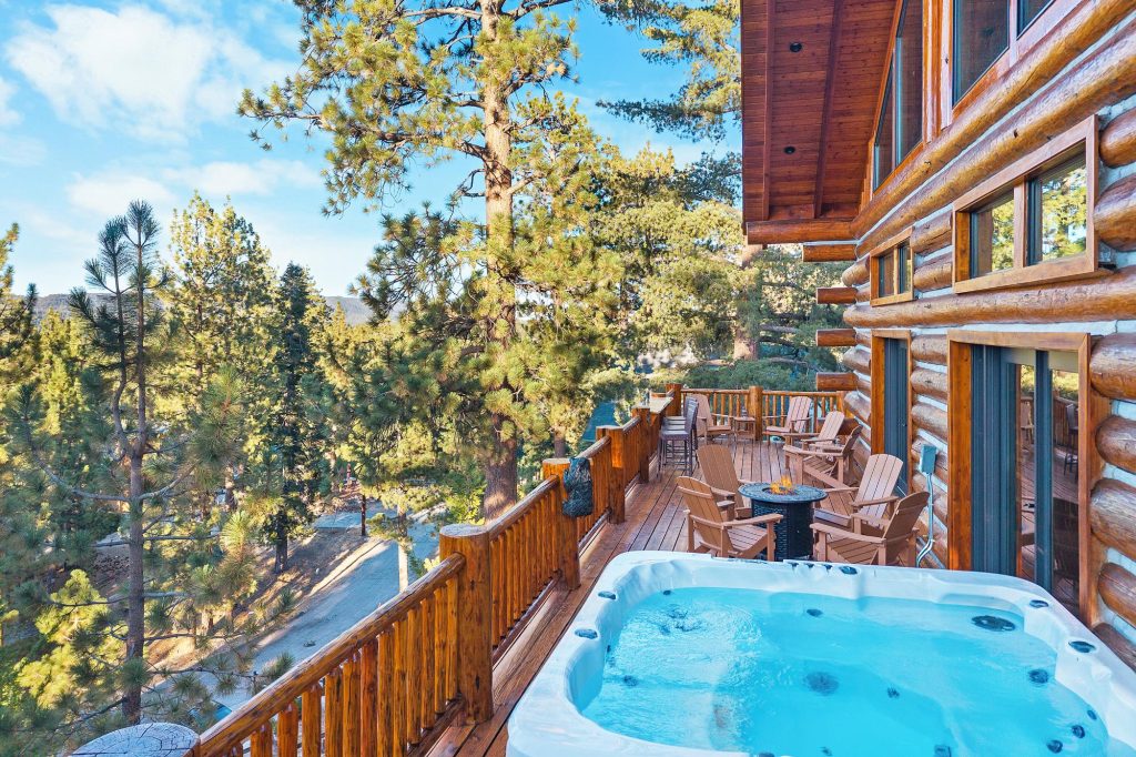 Private cabin balcony with hot tub, wooden deck seating area, and panoramic forest views surrounded by pine trees
