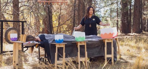 Two clients lying on massage tables outdoors while a practitioner conducts a sound healing session with multiple crystal singing bowls.
