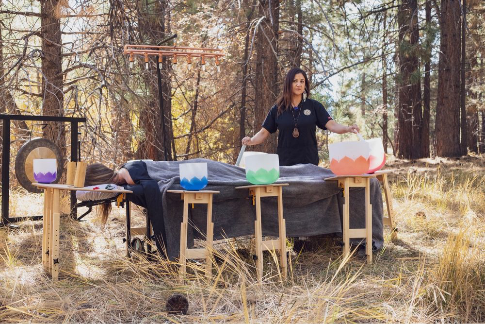 Two clients lying on massage tables outdoors while a practitioner conducts a sound healing session with multiple crystal singing bowls.