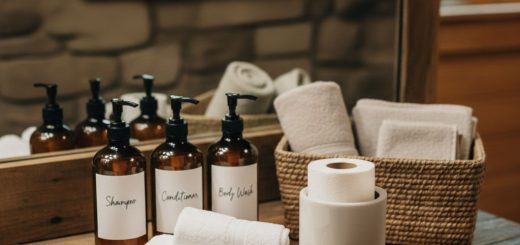 In a bathroom on the counter three dark brown bottles labled shampoo conditioner and body wash, along with towels, soap, and toilet paper on the counter next to a sink with a mirror in back.