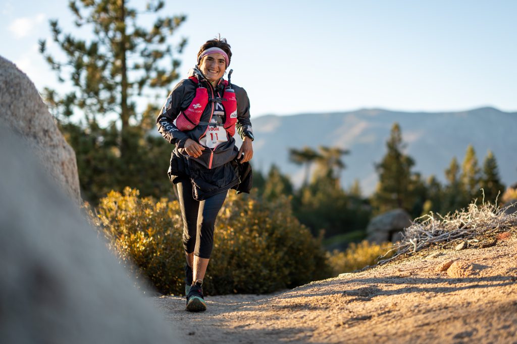 A woman running on a mountain trail in Big Bear, surrounded by lush greenery and rocky terrain.